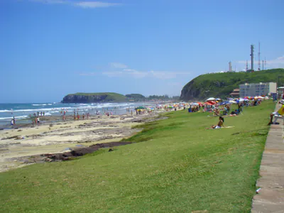 Praia central com gramados naturais, vista para o Morro do Farol e áreas de rochas no mar.