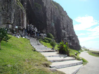 Caminho costeiro entre a Prainha e a Praia da Cal, com pedras, mar e o Oratório Nossa Senhora Aparecida.