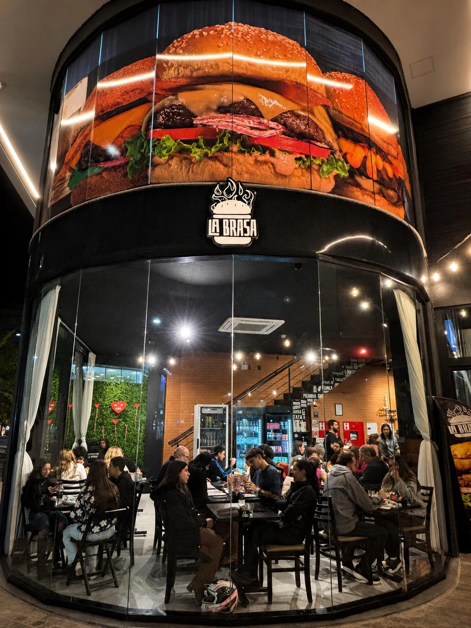 Fachada curva de vidro da La Brasa Burger, com logo e salão em funcionamento