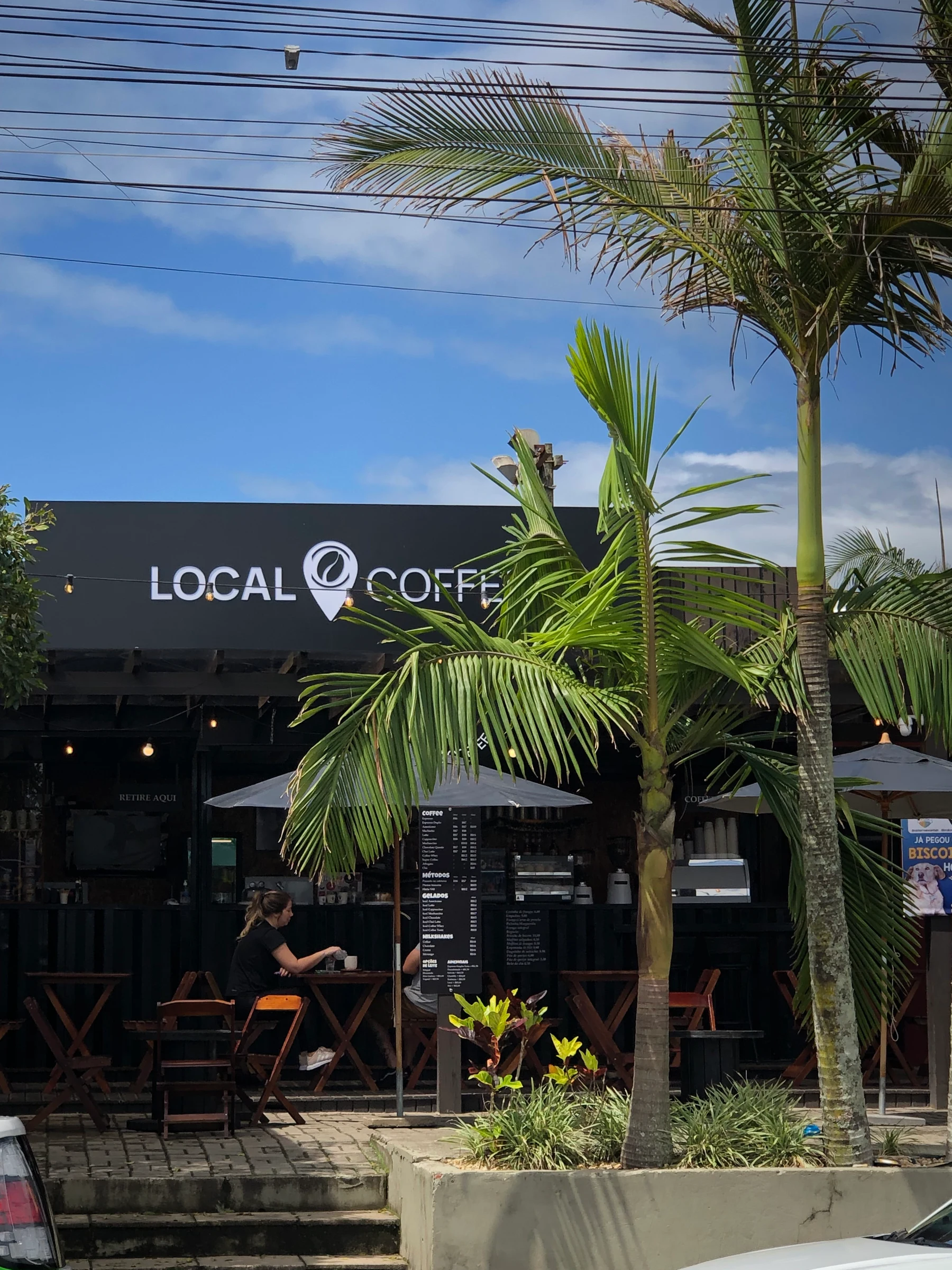 Fachada da Local Coffee com palmeiras, mesas e guarda-sóis
